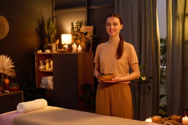 Kafkasyalı genç masözün bej rengi üniformalı uzun at kuyruklu portresi sıcak taşlarla dolu ahşap bir kase tutuyor. Masaj masasını duyuyor musun?