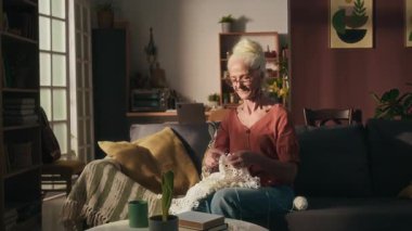 Kameraya gülümseyen gözlüklü ve küpeli beyaz kadın portresi örgü örmek evde kanepede oturan bir hobi.