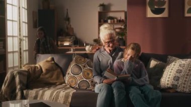 Yaşlı kadın ve kız oturma odasında kitap okuyorlar, mutlu çocuk mutfaktan onlara koşuyor, oturuyor ve büyükannesine sarılıyor.