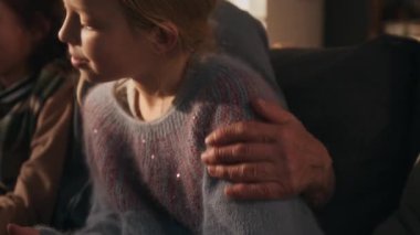 Gen alfa kız mavi süveter giyiyor. Ağabeyi ve büyükannesi birlikte kanepede otururken nazikçe omzunu okşuyor.