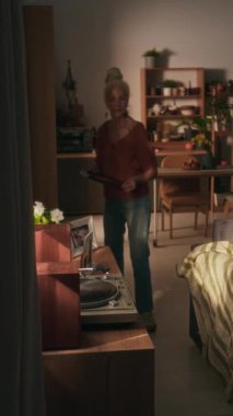 Kafkasyalı, kot pantolon ve kısa kollu bluz giyen yaşlı bir kadının dikey görüntüsü vinil plaklı ahşap bir şifoniyere gidiyor ve onu döner tabağa koyuyor.