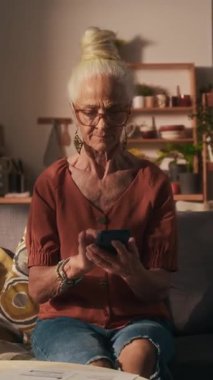 Kafkasyalı yaşlı bir kadının dikey portresi topuz, gözlük ve küpe takıyor ve elinde akıllı telefon ile kameraya poz veriyor.