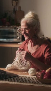 Mutfak masasında dizüstü bilgisayarla video çağrısında akrabalarını selamlayan ve beyaz örgü ören neşeli yaşlı kadının dikey görüntüsü