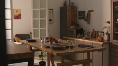 Yemek için hazırlanan ahşap masa, mutfak tezgahı ve duvar dekoru ile aydınlık, rahat mutfak ve yemek alanında çekim yapan kimse yok.