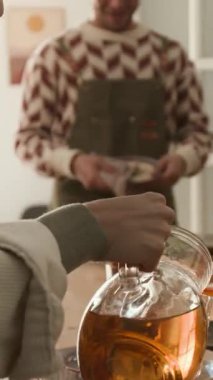 Koyu yeşil önlüklü, gülümseyen bir adamın dikey pozu, arkadaşlarıyla konuşurken ve mutfakta bulaşıkları silerken.