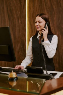 Beyaz tenli genç bir resepsiyonist otel müşterisiyle telefonda konuşurken dikey çekim yapıyor.