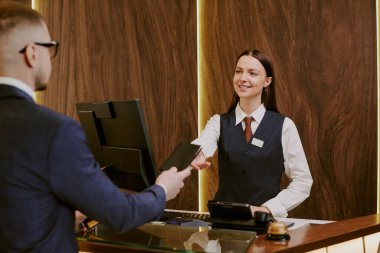 Otel lobisinde masanın yanında duran gülümseyen genç resepsiyoniste pasaport veren tanınmamış bir adam.