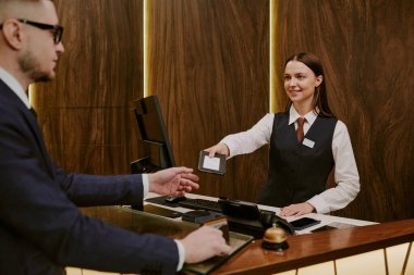 Esmer resepsiyonist, otel misafirlerine pasaport ve oda anahtarı veriyor.