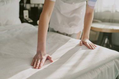 Otel odasındaki çift kişilik yatakta kar beyazı battaniyesini düzelten tanınmayan kadın elleri.