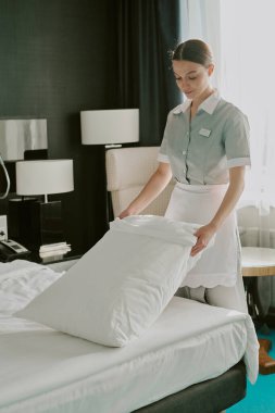 Beyaz üniformalı beyaz oda hizmetçisinin otel odasındaki pamuk beyazı yastık kılıfını düzeltirken dikey görüntüsü.