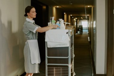 Geniş açılı genç beyaz oda hizmetçisi, açık kapının önünde dururken el arabasının deterjanıyla plastik kutusunu alıyor.