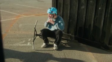 Bisikletle yerde oturan genç bir BMX sporcusu, kask takıyor ve dışarıda antrenman yaparken arkadaşıyla ata biniyor.