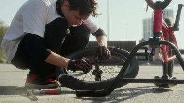 Genç BMX sürücüsü kaldırıma çömelmiş, bisiklet tamir ederken tekerlek üzerinde koli bandıyla kancayı sarıyordu.