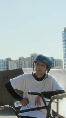 Bisiklet gidonuna yaslanmış, su şişesi tutan ve şehir kaykay parkındaki eğitimden sonra arkadaşıyla konuşan genç BMX sürücüsünün dikey çekimi.