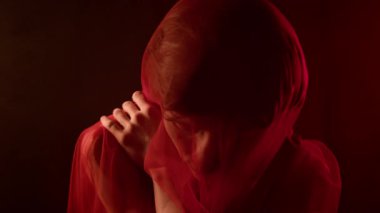 Göğsü kırmızı peçeyle kaplı genç bir adam omuzuna eğik bir şekilde dokunuyor, sonra düşünceli bir şekilde yana bakıyor.