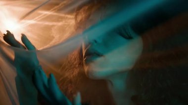 Kıvırcık saçlı, elleri beyaz peçenin altında hareket eden ve gözlerini açıp kameraya bakan rahat genç bir kadının yakın plan portresi.