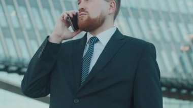 Erkek CEO 'nun telefonda iş konuşurken, modern cam binanın önünde dikilirken düşük açılı görüntüsü.