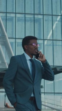 Resmi takım elbiseli Afro-Amerikan bir adamın iş bölgesinde yürürken telefonla konuşmasının dikey çekimi.