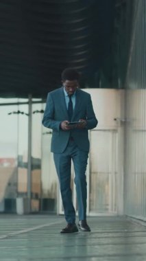 İş için dijital tablet kullanan, resmi takım elbiseli Afro-Amerikan bir adamın şehir merkezindeki ofisine yürürken dikey görüntüsü.