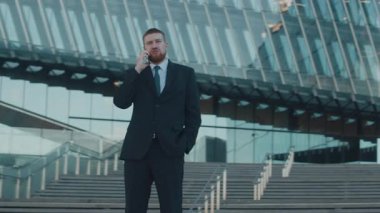 Düşük açılı, koyu takım elbiseli ve kravatlı genç iş adamının, kulağına telefon tutarken, dışarıda dikilirken, parlak, modern cam bina gökyüzünü yansıtırken.