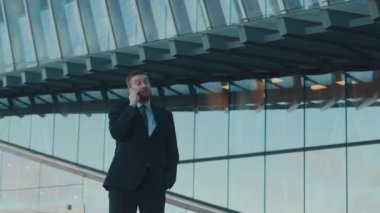 Düşük açılı, keskin giyimli iş adamının cep telefonu konuşması modern kentsel ufuk çizgisini gösteren yansıtıcı cam binanın önünde duruşu.