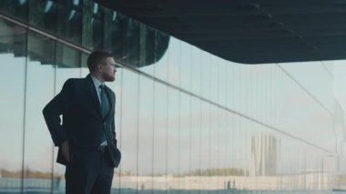 Takım elbiseli sakallı bir adam, telefonuyla mesajlaşıyor, modern cam binanın yanında duruyor, temiz şehir gökyüzünü yansıtıyor.