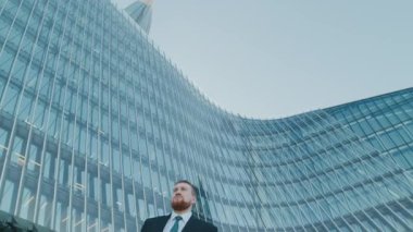 Düşük açılı, keskin giyimli, sakallı bir yöneticinin yükselmeden önce akıllı telefonuyla mesajlaşması, açık mavi gökyüzünü yansıtan kavisli cam bina.