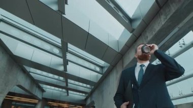 Düşük açılı, takım elbiseli, kahve içen, evrakları taşıyan, modern iş merkezinde yürüyen erkek yöneticinin görüntüsü.