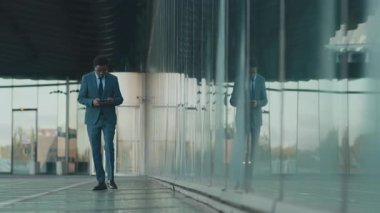 Mavi takım elbiseli Afro-Amerikan adam, modern binanın camına yansıdı, yürüdü, elindeki dijital tablete odaklandı, geniş kentsel iş bölgesinde.