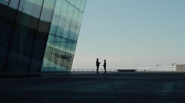 Birbirlerine bakan tanınmayan iki işadamı, yansıtıcı modern cam bina tarafından çerçevelenmiş geniş asfalt plazada çalışmaktan bahsediyorlar.