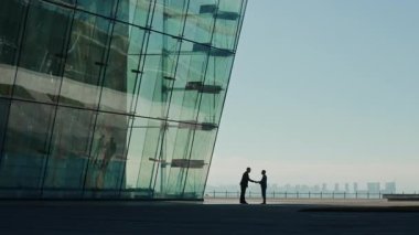 Tanımlanamayan iki insan cam bina önünde el sıkışıyor ve birlikte yürüyorlar, siluetleri şehir manzarasına karşı yansıyor.