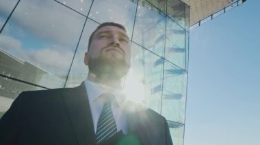 Düşük açılı, koyu renk takım elbiseli, sakallı bir adamın modern cam binaya dalgın dalgın bakarken çekilmiş portresi.