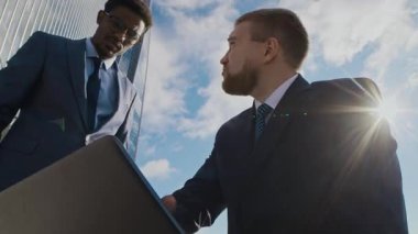 Afrika kökenli Amerikalı bir adamın resmi iki parçalı takım elbisesiyle iş yerindeki uzun ve cam bina dışında meslektaşına dizüstü bilgisayarda yardım ederken düşük açılı görüntüsü.