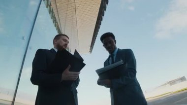 Takım elbiseli iki adam ellerinde belgelerle cam bina dışında konuşuyorlardı. Yansımaları modern mimaride yansıtılıyordu.