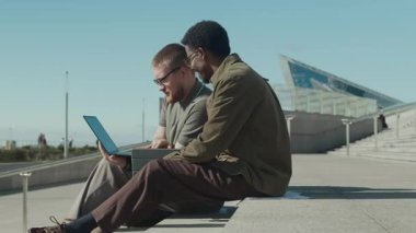 Kısa kollu gömlekli sakallı adam ve ceketli kıvırcık saçlı Afro-Amerikan adam dışarıda beton merdivenlerde otururken laptopta iş tartışıyorlar.