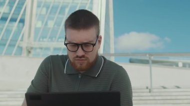 Laptopta çalışan, gözlüklerini ayarlayan, kameraya bakan ve iş yerindeki beton merdivenlerde oturan sakallı adamın portresi.