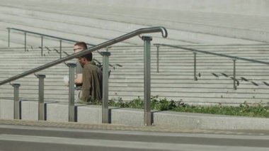 Parlak bir günde modern cam binanın önünde metal parmaklıklı beton merdivenlerle yürüyen iki adam.