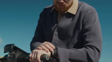 Gözlüklü genç sakallı adam duraklıyor. Elektrikli scooter kullanıyor ve güneşli bir günde parkta saati kontrol ediyor.