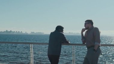 Güvertede güneşli bir günde korkuluklara yaslanmış, parıldayan suya tepeden bakan iki adam gündelik bir konuşma yapıyor.