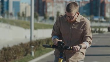 Kahverengi gömlekli sakallı bir adam asfalt yolda elektrikli scooter ayarlıyor. Çimenli alana ve şehir manzarasına bakıyor.