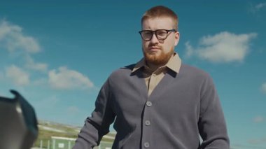 Bej gömlekli ve hırkalı sakallı bir adam akıllı telefonunu elektrikli scooter 'a takıyor ve güneşli bir günde parka gidiyor.