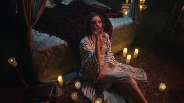 Solgun vampir kadının mum ışığıyla çevrili, rahat ve derin düşüncelere dalmış bir şekilde kan içtiği yüksek açılı bir çekim.