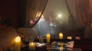 Sönük odasında dans eden vampir kadın mum ışığında ısınıyor ve kısmen perdeli yatak ve dağınık nesnelerle örtülmüş.