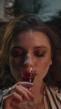 Büyüleyici makyajlı yırtıcı vampir kadının küçük bir bardak kanın tadını çıkarırken dikey açıyla çekilen görüntüsü.
