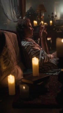 Çizgili bornozlu vampir kadının dikey resmi kitapların arasında dinleniyor, loş ışıkta bir bardak kan tutuyor, mum ışığıyla dolu gotik bir oda.