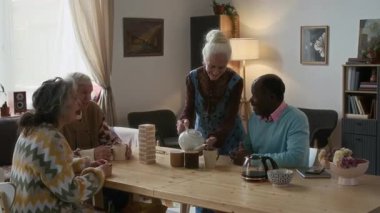 Bir grup yaşlı, çok ırklı arkadaş ahşap masanın etrafında oturur, çaydan keyif alır, sohbet eder ve huzurevinin rahat oturma odasında oyun oynarlar.