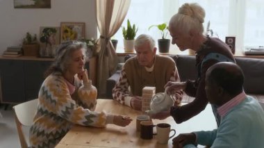 Huzurevinin rahat odasında, ahşap masada oyun oynayan bir grup arkadaşa çay servisi yapan kıdemli bir kadın.