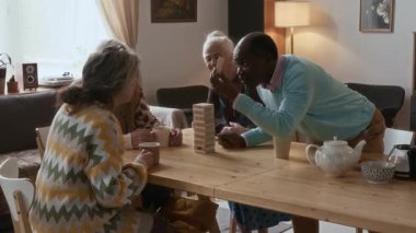 Çok ırklı yaşlı arkadaşlar, rahat oturma odasındaki ahşap masanın etrafında toplanmış, huzur evinde boş zamanlarını geçirirken bloklama oyunu oynuyorlardı.