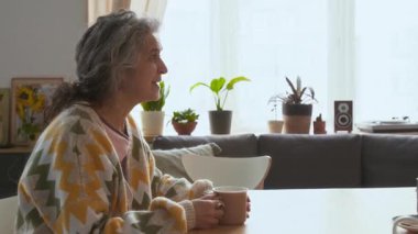 Yaşlı gri saçlı kadın rahat oturma odasında oturuyor, fincandan çay içiyor ve huzurevindeki arkadaşıyla konuşuyor.