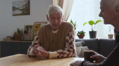 Yaşlı adam oturma odasında masada oturuyor, kağıt karıştırıyor ve kreşte arkadaşlarıyla sohbet ediyor.